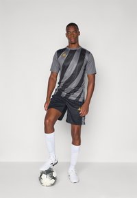 Grå treningstrøye med diagonale svarte striper, gulllogo; svarte shortser; hvite knelange sokker; står på en svart-hvitt fotball.