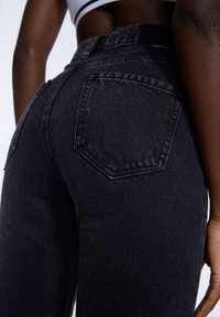 Un primer plano de una persona que lleva jeans de mezclilla negros de tiro alto con un bolsillo trasero y una camiseta corta blanca.