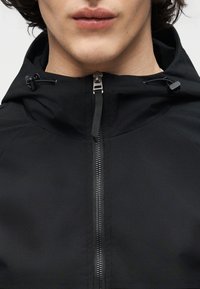 Νεαρός φορεί μαύρο μπουφάν με φερμουάρ και ρυθμιζόμενη κουκούλα, με ορατό φερμουάρ και κορδόνια στο λαιμό.