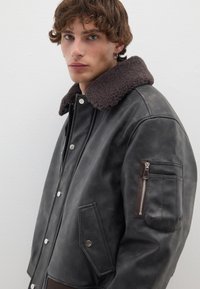 Giacca di pelle nera con colletto in shearling marrone, chiusura frontale a scatto, tasche laterali e tasca con zip sulla manica sinistra.