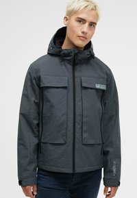 Szara wodoodporna kurtka z zamkiem błyskawicznym, regulowanym kapturem, dwiema dużymi kieszeniami na piersi oraz detalem logo na lewej rękawie. Gładka struktura.