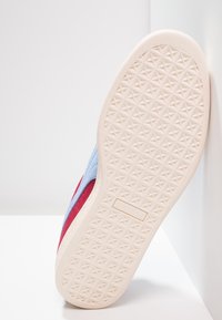 Sneaker med en överdel i röd och blå mocka, med en texturerad krämfärgad gummisula med geometriskt mönster. Logotyp präglad på sidan.