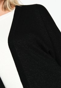 Svart, texturerad cardigan med en lös passform och öppen front, som hänger över en vit ribbad topp. Har subtil glans genom hela tyget.