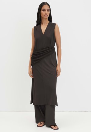 Frau trägt ein ärmelloses schwarzes Kleid mit V-Ausschnitt und geflochtenem Gürtel-Detail, kombiniert mit weiten schwarzen Hosen und schwarzen Sandalen.