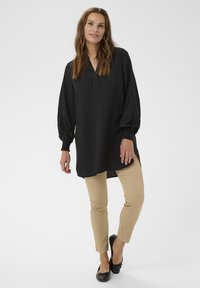 Žena stojí oblečená v voľnej čiernej blúzke s dlhým rukávom, béžových štíhlych nohaviciach a čiernych baletných topánkach pred jednoduchým svetlým pozadím.