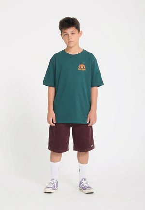 Un garçon est debout, faisant face à l'avant, portant un T-shirt Volcom vert, un short bordeaux, des chaussettes blanches et des baskets violettes sur un fond blanc.