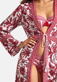 Rode kimono met bloemenpatroon en brede mouwen, op een witte achtergrond, vastgemaakt met een witte koord, waaronder een bijpassende bikini zichtbaar is.