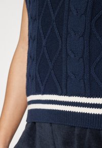 Pull sans manches en tricot bleu marine avec des motifs de câbles texturés, présentant une bande horizontale blanche à l'ourlet. Tissu doux.