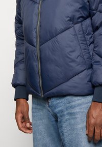 Navyblauwe pufferjack met quilted patronen, een rits aan de voorkant en ribgebreide manchetten. Gecombineerd met lichtblauwe jeans. Zachte textuur.