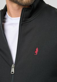 Svart hettejakke med høy krage, med en liten rød brodert figur på venstre bryst, båret over en hvit t-skjorte.