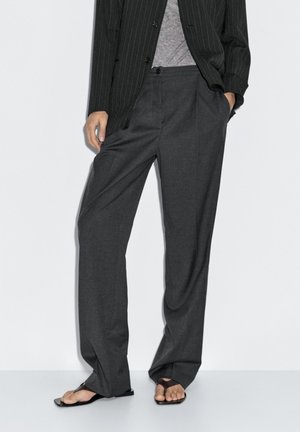Pantaloni grigi scuri, larghi e realizzati in tessuto leggero, con parte anteriore piatta e tasche laterali. Abbinati a sandali casual.