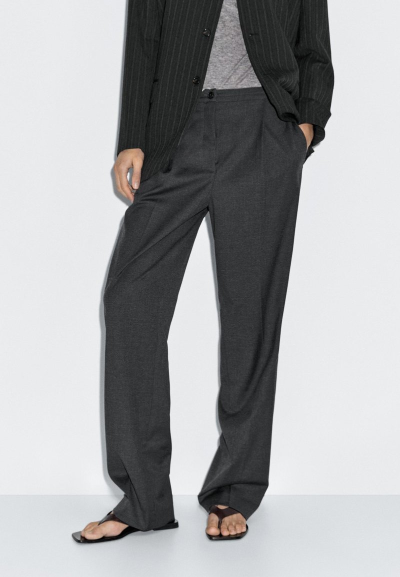 Pantaloni grigi scuri, larghi e realizzati in tessuto leggero, con parte anteriore piatta e tasche laterali. Abbinati a sandali casual.