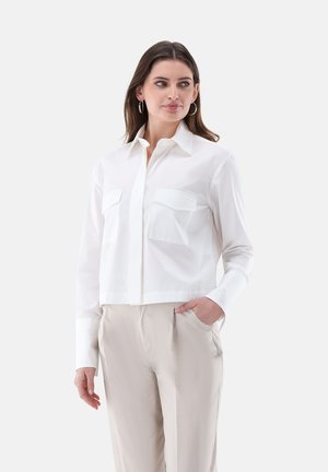 Donna che indossa una camicia bianca con bottoni e grandi tasche frontali e pantaloni beige, in piedi con una mano in tasca davanti a uno sfondo semplice.