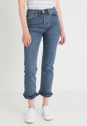 Woman wearing blue denim jeans with ruffled hems, white sneakers, and a striped t-shirt, standing against a plain light background.