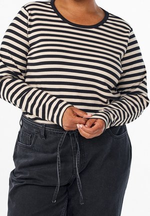 Persoon draagt een zwart-wit gestreept shirt met lange mouwen en zwarte hooggetailleerde broek, gestrikt in de taille.