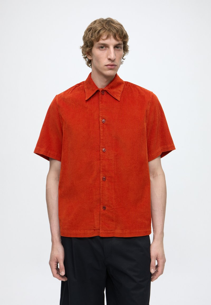 Jeune homme portant une chemise en velours côtelé rouge à manches courtes boutonnée et un pantalon noir, debout devant un fond clair uni.