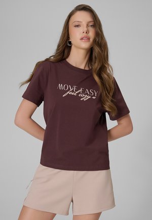 Kobieta z długimi falującymi włosami, ubrana w ciemnobrązowy T-shirt z napisem "Move Easy feel cozy" oraz jasnobeżowe krótkie spodenki, stoi na neutralnym tle.