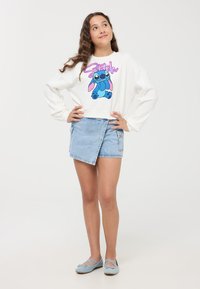 Sweatshirt bianco corto con stampa di un personaggio dei cartoni animati blu, abbinato a una mini gonna di jeans chiari con design a portafoglio e tasche visibili.