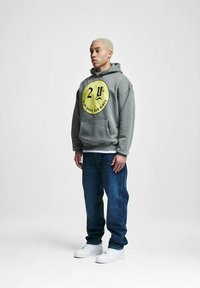 Grå hettegenser med et gult sirkulært grafisk motiv, med tallene "24" og teksten "F**k dine dårlige vibber." Bært med blå jeans og hvite sneakers.
