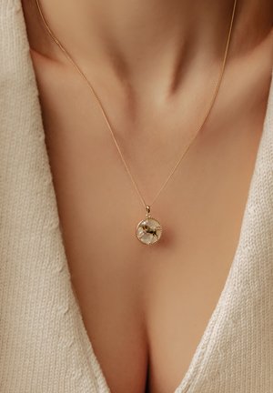 Halskette aus Goldfarbe mit einem runden Anhänger auf weißem Hintergrund und einer detaillierten goldenen Figur. Feine Kette, minimalistisches Design.