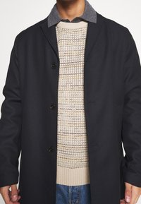 Cappotto scuro a trama con rever a punta, bottoni neri, indossato sopra un maglione leggero a maglia con un motivo beige e marrone.