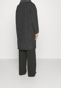 Abrigo acolchado negro de longitud media con patrones circulares, que presenta un cuello gris y puños abotonados, combinado con pantalones oscuros holgados.