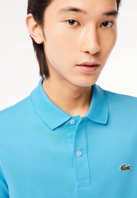 Camisa polo turquesa en tejido texturizado, con cuello, dos botones y un logo de cocodrilo bordado en verde en el lado izquierdo del pecho.