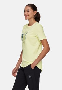 Lichtgele T-shirt met een ronde hals en korte mouwen; heeft een grafisch ontwerp in blauw en groen. Draagt met donkere broeken en zijzakken.