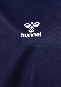 Marinblått texturerat tyg med en vit "hummel"-logotyp med en stiliserad bi-design, placerad tydligt på vänstra sidan.