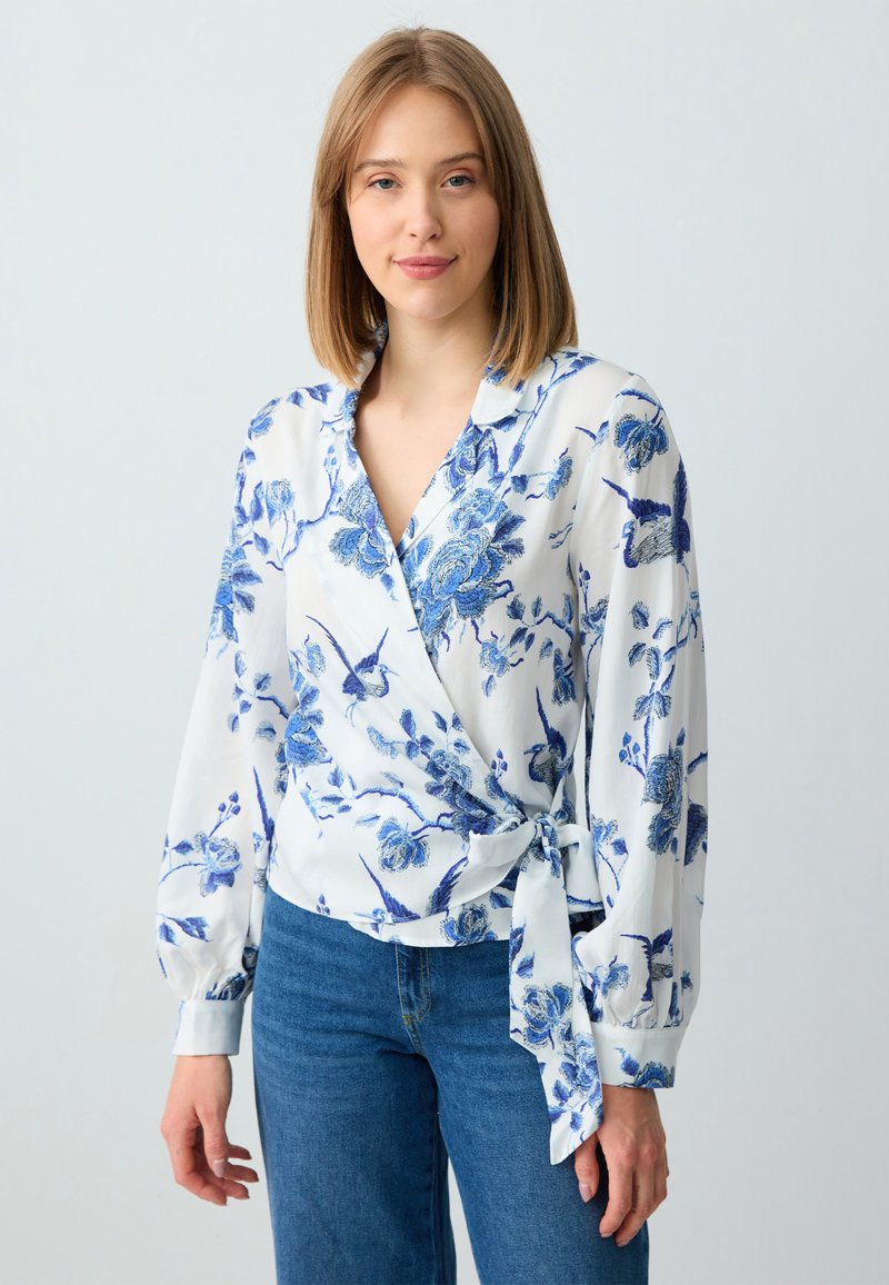 Weiße Wickelbluse mit blauem Blumenmuster; hat lange Ärmel und eine gebundene Taillenpartie. Weicher Stoff mit fließender Textur. Kombiniert mit blauen Jeans.