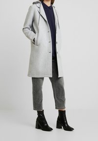 Grijze capuchonjas gemaakt van zachte stof, met een knopenfront, zijzakken en een relaxed fit. Geselecteerd met donkergrijze jeans en zwarte hoge laarzen.