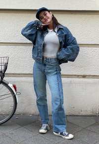 Jachetă din denim, bluză scurtă cu dungi albe, blugi deschiși cu talie înaltă, și șapcă bleumarin, asociate cu adidași bleumarin și albi. Fundalul include o bicicletă.