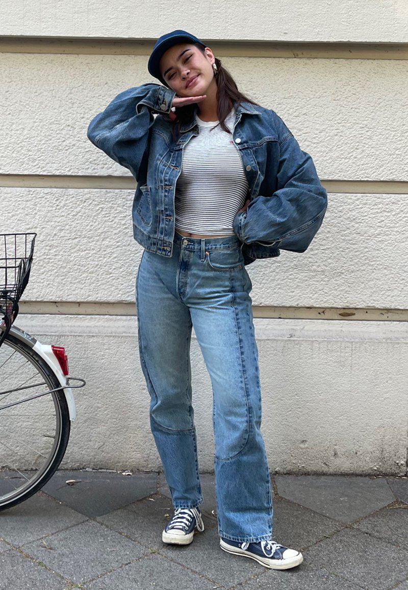 Denimjakke, hvitstripet crop topp, lyseblå høy-liv jeans, og marineblå caps, kombinert med marineblå og hvite joggesko. Bakgrunnen inkluderer en sykkel.