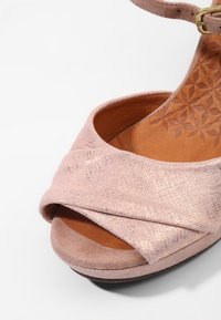 Gros plan sur une sandale à talon ouverte en daim rose avec des lanières croisées, une semelle intérieure beige embossée de motifs floraux et une bride réglable à la cheville avec une boucle dorée.
