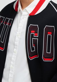 Zwarte bomberjack met een rood en wit gestreepte kraag, met grote "HUGO" letters in een gestructureerd ontwerp op de voorkant. Draagt over een wit shirt.