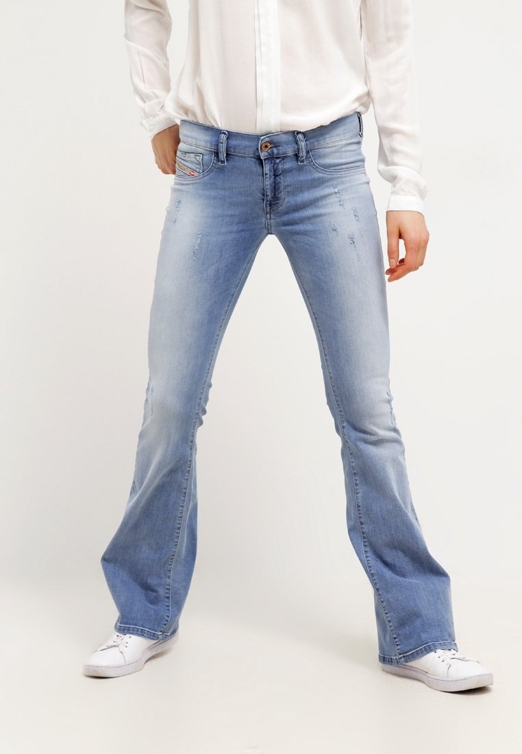 Personne portant un jean évasé bleu clair délavé, des baskets blanches et une chemise blanche à manches longues, debout jambes écartées devant un fond uni.