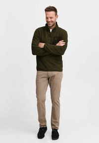 Pull-over en polaire vert olive avec un col haut et une poche zippée sur la poitrine, associé à un pantalon beige clair et des chaussures noires.