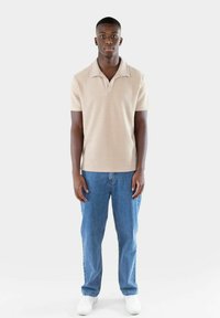 Beige ribbad polo skjorta med bred krage, matchad med ljusblå raka jeans och vita sneakers. Enkelt design, framifrån.