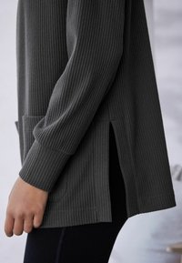 Haut long à manches raglan en maille côtelée gris foncé avec fentes latérales et poches, présentant un tissu texturé et une coupe décontractée, contrastant avec des leggings noirs unis.