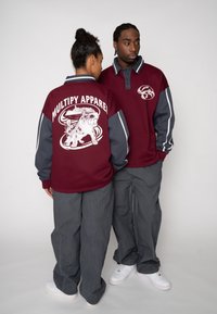 Bordeauxroter Pullover mit grauen Ärmeln, mit dem Schriftzug "MULTIPY APPAREL" und einem grafischen Design auf der Rückseite. Graue Hose und weiße Turnschuhe.