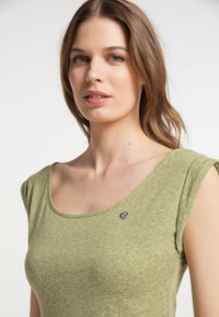 Maslinasto zelena bluza kratkih rukava s okruglim izrezom i suptilnom teksturom. Ima mali crni logo od šivanja blizu ovratnika.