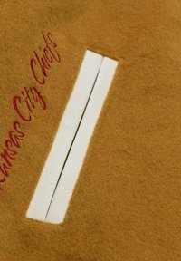 Brown wool fabric with a vertical white leather strip and red embroidered text reading "Kansas City Chiefs" beside it.