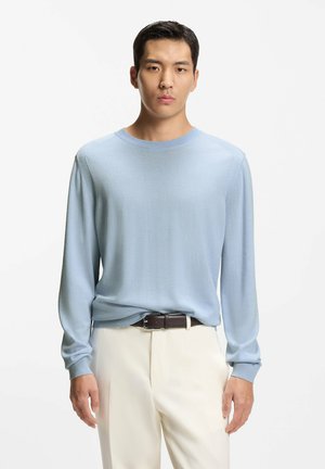 Jeune homme portant un pull bleu clair à manches longues, rentré dans un pantalon crème avec une ceinture noire, debout devant un fond blanc uni.