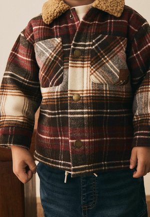 Enfant portant une veste à carreaux rouge, marron et crème avec de grandes poches avant et un col en polaire beige, assortie à un jean bleu.