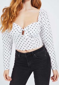 Haut court blanc à pois avec un col en cœur, des manches longues et un détail de nœud à l'avant, associé à un jean noir taille haute.