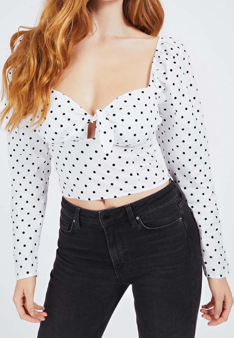 Haut court blanc à pois avec un col en cœur, des manches longues et un détail de nœud à l'avant, associé à un jean noir taille haute.