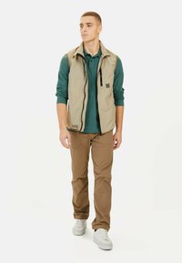 Beige ærmeløs vest over en mørkegrøn langærmet skjorte, parret med brune bukser og hvide sneakers. Har en lynlåslomme og letvægtsmateriale.