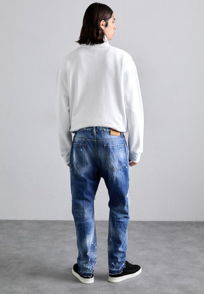 Vit sweatshirt, blå slitna jeans med färgfläckar, två bakfickor och en läderlapp. Svarta sneakers fulländar looken.
