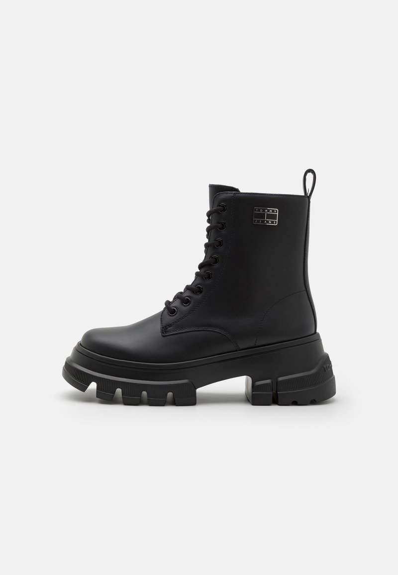 Botines negros con cordones hechos de cuero sintético. Presentan una suela gruesa y texturizada, y una lengüeta en la parte trasera. Sutil branding en el costado.