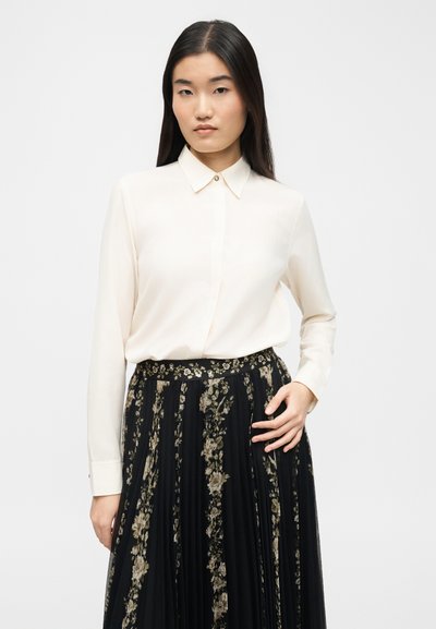 Γυναίκα με μακριά σκούρα μαλλιά φοράει κρεμ πουκάμισο με κουμπιά και μαύρη φουστάνια με λουλούδια και πιέτες, στέκεται μπροστά σε απλό φωτεινό φόντο.